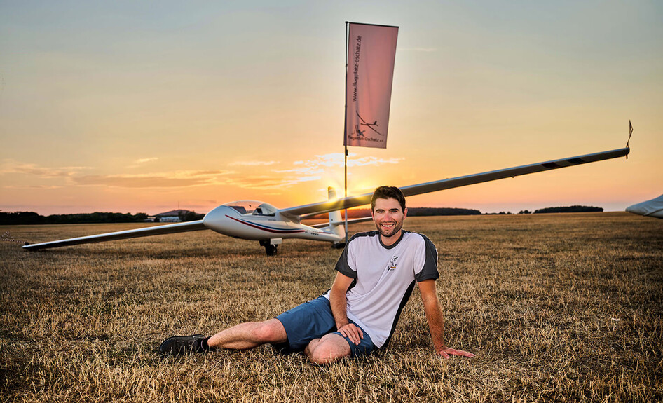 Person sitzt vor einem Segelflugzeug auf einer Wiese bei Sonnenuntergang